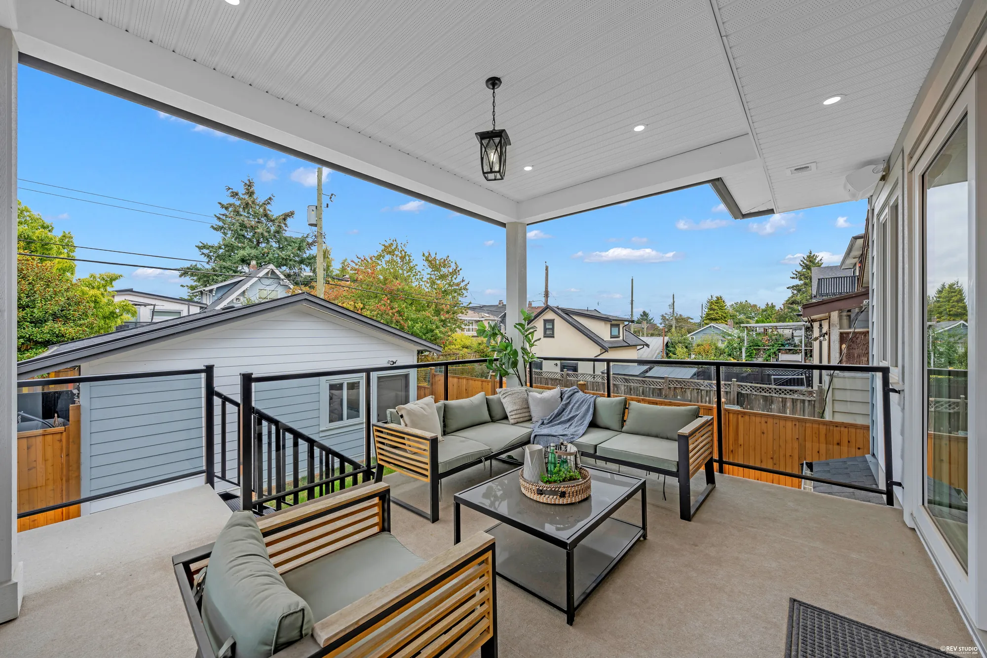 The covered balcony off the living room: a white ceiling with a lantern, black railings, an outdoor sofa and chairs, the yard and the lane beyond