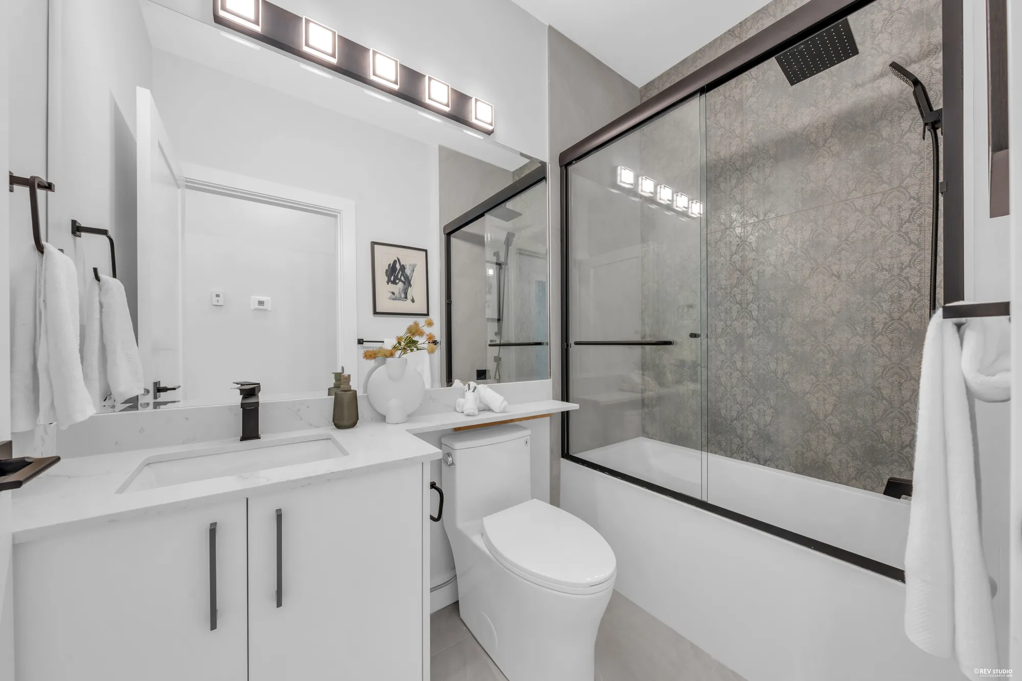 A bathroom with a tub behind a sliding glass screen, a white vanity under a mirror, a row of lights