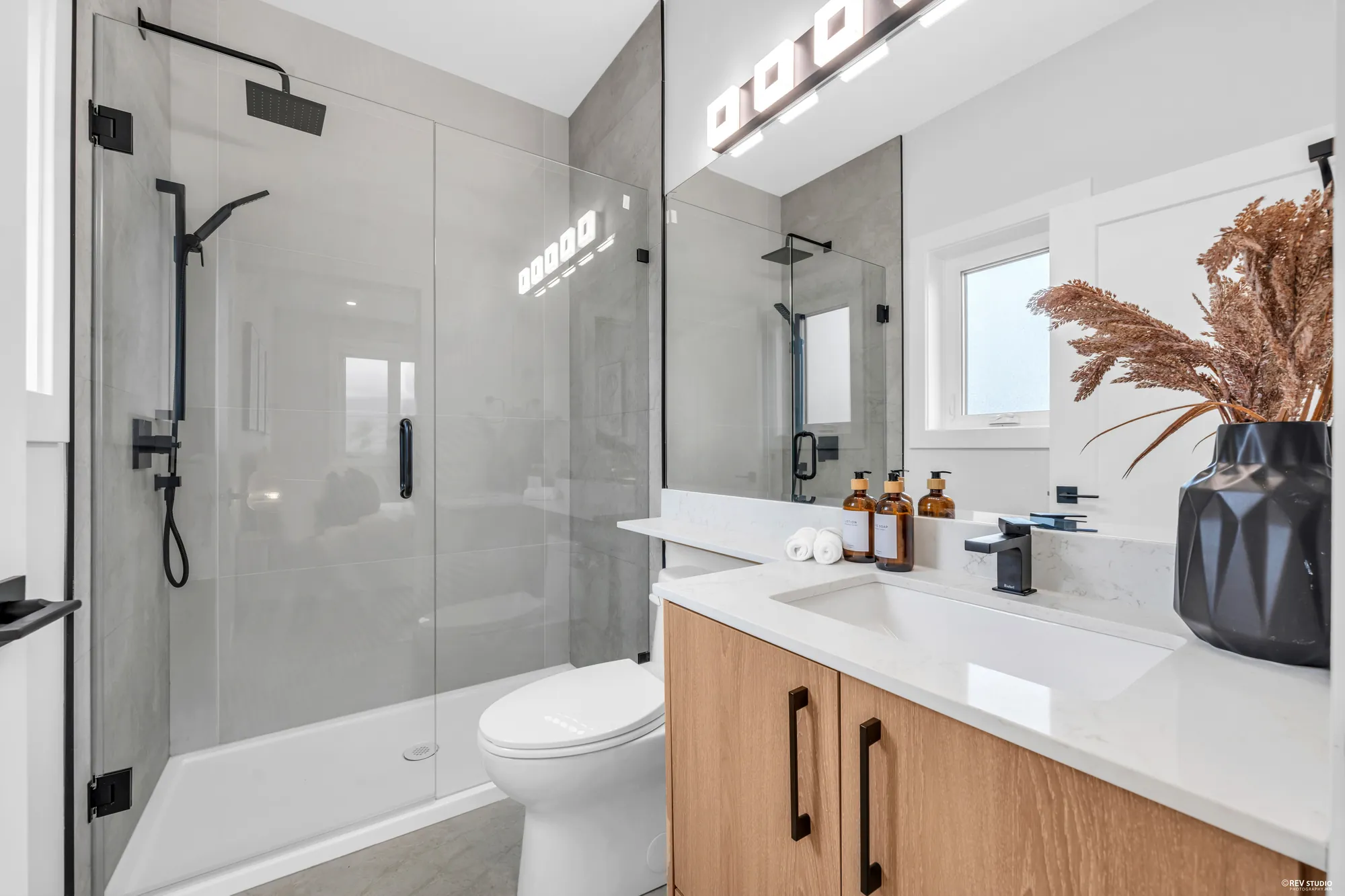 A bathroom with a glass shower in grey tile, an oak vanity under a mirror, a window