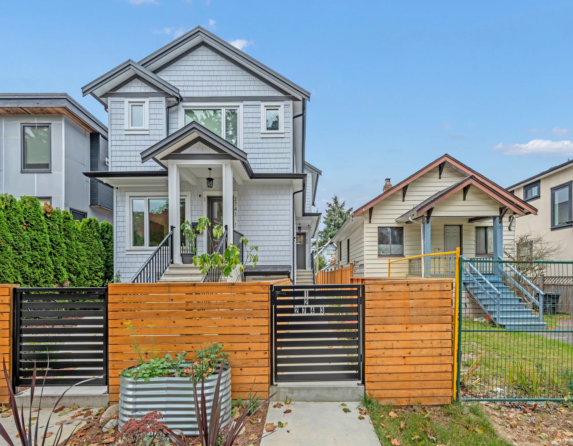 The front of the house in daylight: three levels of white shingle and board with black trim, the black gate in the cedar fence with the number on it, two steel planters, the older neighbour beyond