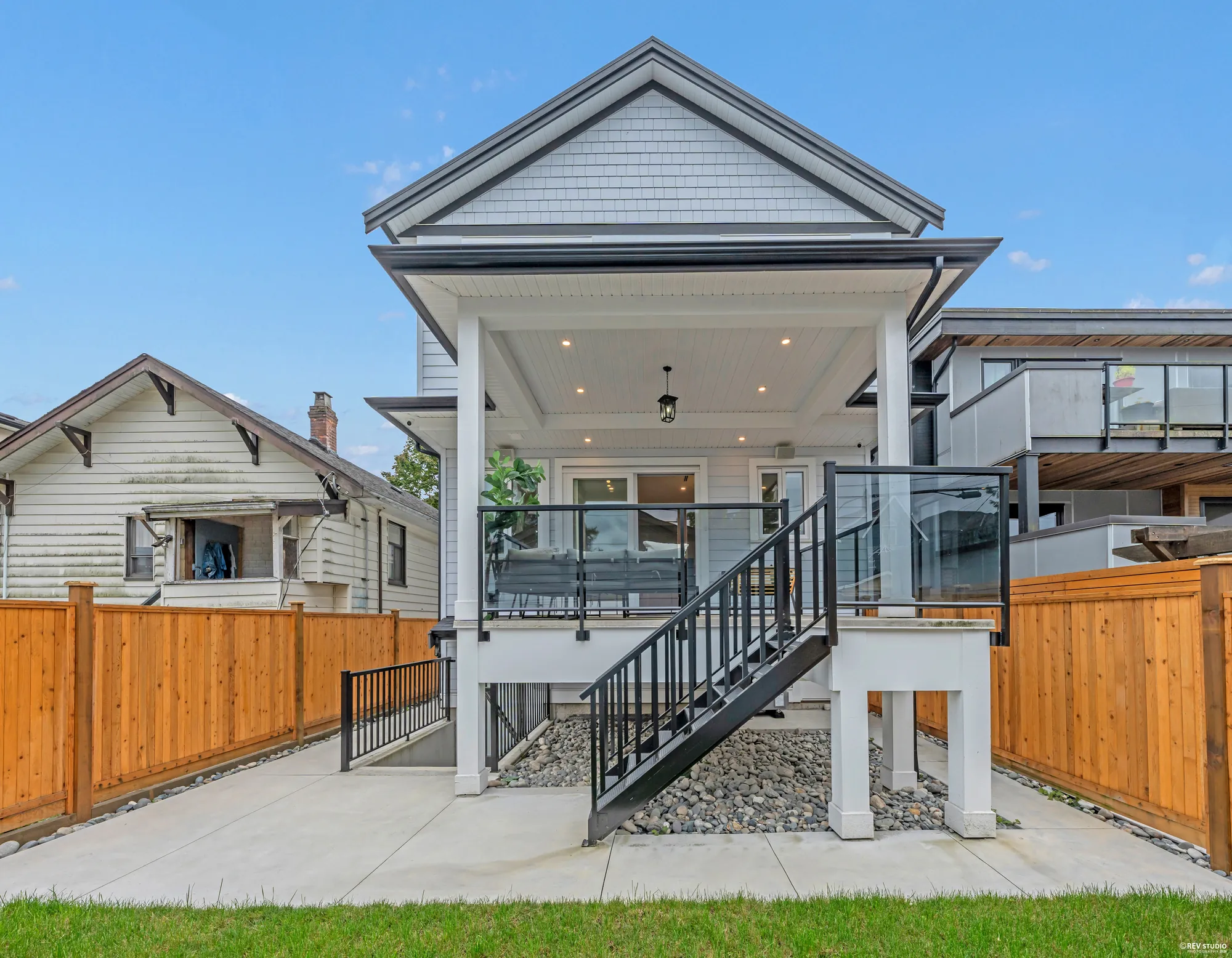 The back of the house straight on: the covered balcony with its black railings and a glass panel, the stair down to a path, the cedar fence both sides, turf in front