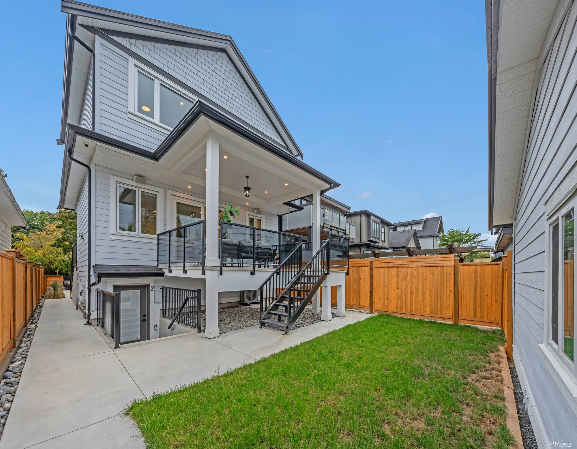The back of the house from the corner of the yard: the covered balcony under the gable, the stair down, the turf, the neighbour's older house beyond the fence